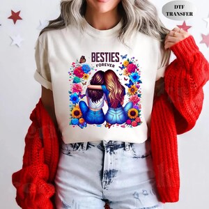 May include: Cream-colored t-shirt featuring a graphic of two embracing figures with the text "BESTIES FOREVER." The design is surrounded by colorful flowers and butterflies. The shirt is a casual wear item.