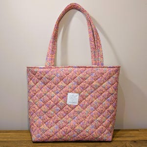 Può includere: Una borsa tote trapuntata con un motivo floreale nei toni del rosa, viola e giallo. La borsa ha due manici e un motivo a rombi. Un'etichetta bianca con testo è cucita sul davanti.