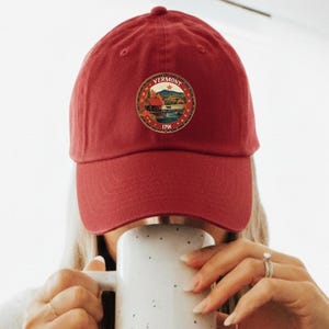 May include: A red baseball cap with a Vermont emblem is worn by a person holding a white speckled mug with a brown base. The mug is filled with a beverage. The person is wearing a gray t-shirt and gray sweatpants.