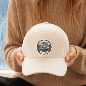 May include: A cream-colored baseball cap featuring a circular emblem with the text "CONNECTICUT" and "1788". The emblem includes a depiction of a ship, trees, and a lighthouse. The cap is held in front of a snowy winter scene.