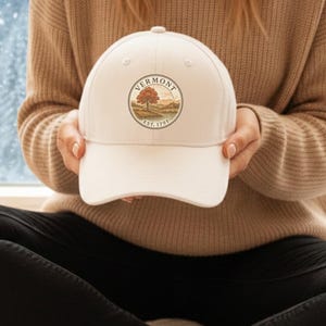 May include: A white baseball cap with a circular design featuring a tree and the text "VERMONT EST. 1791". The cap is held in front of a snowy winter landscape.
