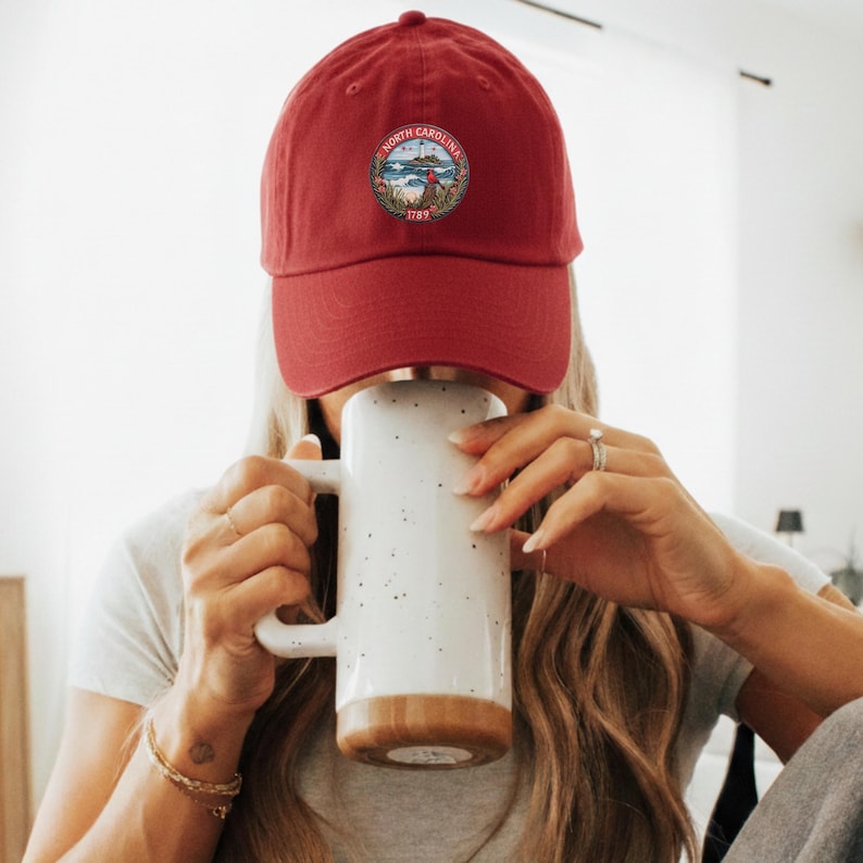 Op de afbeelding: Rode baseballcap met een North Carolina-embleem. Een persoon houdt een grote witte mok met een bruine basis vast. De mok heeft een gespikkeld ontwerp.
