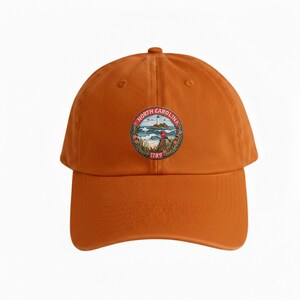Op de afbeelding: Oranje baseballpet met een cirkelvormig embleem met de tekst "North Carolina" en het jaar "1789". Het embleem toont een vuurtoren, oceaangolven en een rode vogel. De pet heeft een gebogen klep en een verstelbare band.
