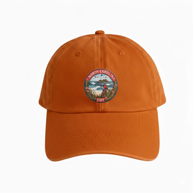 Op de afbeelding: Oranje baseballpet met een cirkelvormig embleem met de tekst "North Carolina" en het jaar "1789". Het embleem toont een vuurtoren, oceaangolven en een rode vogel. De pet heeft een gebogen klep en een verstelbare band.