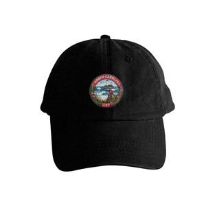 Op de afbeelding: Zwarte baseballcap met een cirkelvormig embleem met een vuurtoren, oceaangolven en de tekst "North Carolina 1789". Het embleem is omlijst door een rood-witte rand met bloemmotieven. De cap heeft een gebogen klep.