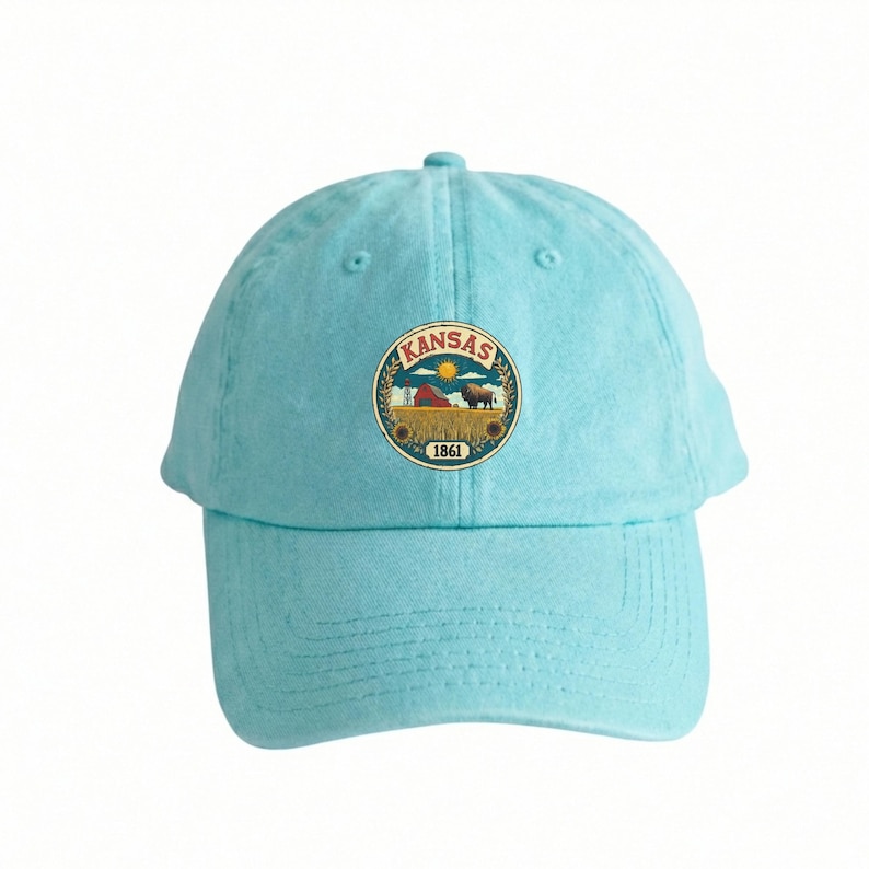 K&ouml;nnte beinhalten: Hellblaue Baseballkappe mit einem kreisf&ouml;rmigen Emblem mit dem Wort "KANSAS" und einer l&auml;ndlichen Szene. Das Emblem zeigt eine Scheune, ein Weizenfeld, einen Bison und das Jahr 1861. Die Kappe ist aus weichem Stoff.
