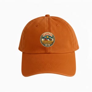 K&ouml;nnte beinhalten: Gebrannte orangefarbene Baseballkappe mit einem kreisf&ouml;rmigen Emblem. Das Emblem zeigt das Wort "Kansas" und eine Farm-Szene mit Scheune, Bison und Sonnenblumen. Das Jahr "1861" befindet sich am unteren Rand des Emblems. Klassischer Stil.