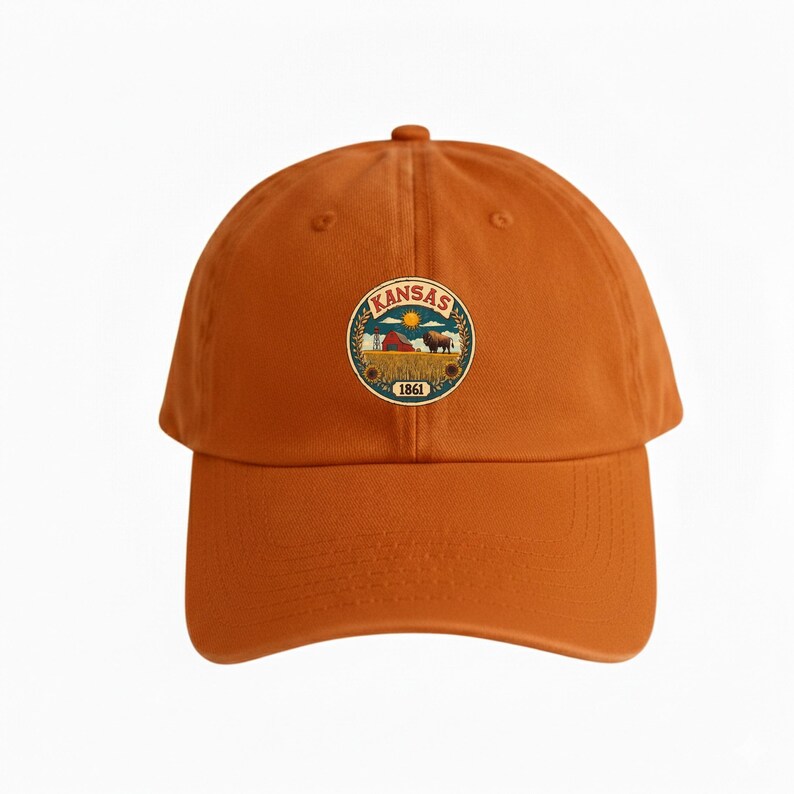 K&ouml;nnte beinhalten: Gebrannte orangefarbene Baseballkappe mit einem kreisf&ouml;rmigen Emblem. Das Emblem zeigt das Wort "Kansas" und eine Farm-Szene mit Scheune, Bison und Sonnenblumen. Das Jahr "1861" befindet sich am unteren Rand des Emblems. Klassischer Stil.