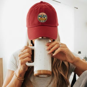 K&ouml;nnte beinhalten: Burgunderfarbene Baseballkappe mit einem runden Aufn&auml;her mit dem Wort "Kansas" und einem Landschaftsdesign. Die Kappe wird von einer Person getragen, die einen wei&szlig;en Becher mit braunem Boden h&auml;lt.