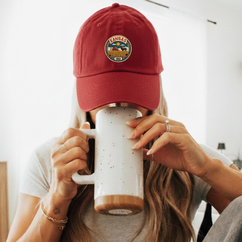 K&ouml;nnte beinhalten: Burgunderfarbene Baseballkappe mit einem runden Aufn&auml;her mit dem Wort "Kansas" und einem Landschaftsdesign. Die Kappe wird von einer Person getragen, die einen wei&szlig;en Becher mit braunem Boden h&auml;lt.
