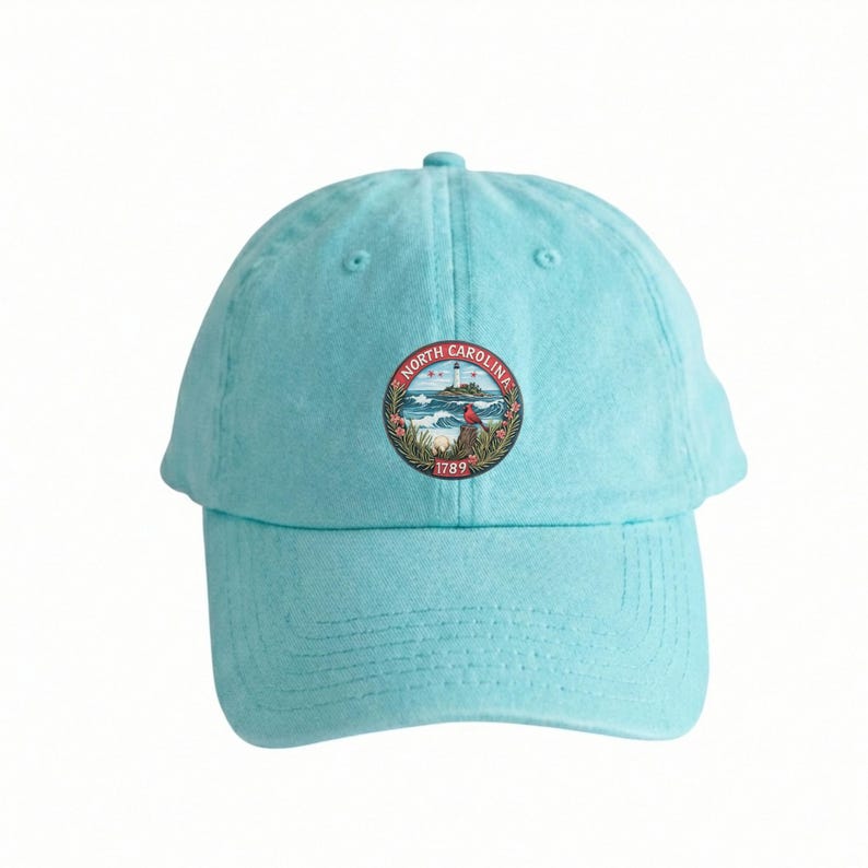 Op de afbeelding: Lichtblauwe baseballcap met een cirkelvormig embleem met de tekst "North Carolina" en het jaar "1789". Het embleem toont een vuurtoren, golven en een vogel. De cap is gemaakt van een zachte stof.