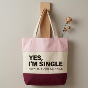 May include: A canvas tote bag with a pink, cream, and burgundy color block design. The bag has cream-colored handles and features the text "YES, I'M SINGLE NOW IS YOUR CHANCE" in black. A dried rose is placed inside the bag.