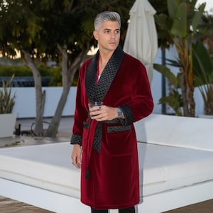 Op de afbeelding: Een bordeauxrode fluwelen badjas met zwarte gewatteerde revers, manchetten en ceintuur. De badjas heeft een sjaalkraag en zakken. Het model draagt een horloge en houdt een glas vast. Een luxueus kledingstuk.