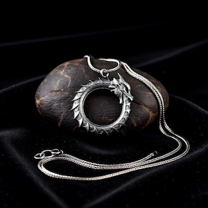 May include: A silver-colored dragon pendant necklace. The pendant is a circular design with a dragon head and tail forming a complete circle. The necklace chain is a thin, woven style. The pendant is resting on a dark, textured stone against a black background.