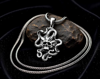 May include: A silver-colored pendant necklace featuring a skull design surrounded by octopus tentacles. The pendant is suspended from a matching silver chain. The necklace is displayed on a dark, textured surface.