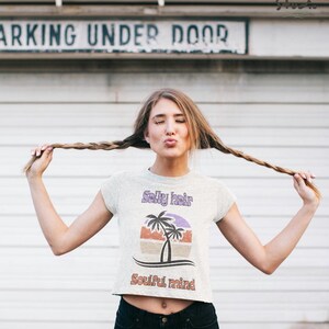 May include: A light gray crop top with the text "Salty hair Soulful mind" and a graphic of palm trees. The model is holding her long braided hair up. The background is a white garage door.