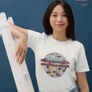 Puede incluir: Camiseta blanca con un diseño gráfico de bola de discoteca colorida y el texto "Always enough, never too much". La persona lleva vaqueros azules. El fondo es azul oscuro.