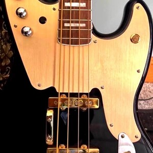 May include: A black electric bass guitar featuring a brushed gold pickguard, four strings, a dark wood fretboard with rectangular inlays, gold-colored bridge pickups, and four silver control knobs mounted on the upper horn of the body.