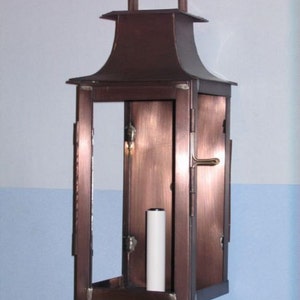 May include: A copper-colored wall-mounted lantern with a glass front and a single light bulb inside. The lantern has a square shape with a decorative top.