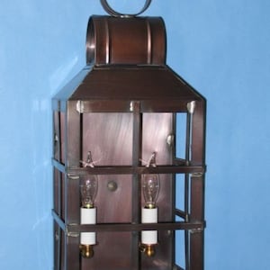 May include: A copper and black metal wall sconce with two light bulbs. The sconce has a rectangular shape with a decorative top.