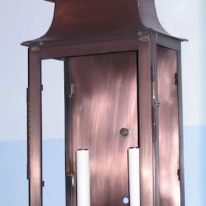 May include: A copper wall-mounted lantern with a rectangular glass panel and two light bulbs. The lantern has a decorative top with a handle.