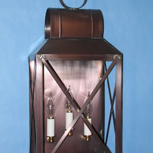 May include: A copper wall lantern with a rectangular frame and a decorative top. The lantern has three light bulbs and a criss-cross pattern on the front.