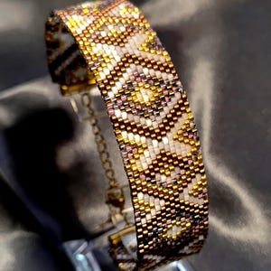 May include: A beaded bracelet with a geometric pattern in gold, white, and brown tones. The bracelet is made of small beads and has a gold-colored chain clasp. The design features a repeating pattern of diamonds and zigzags.