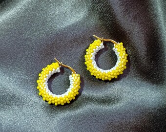 May include: A pair of beaded hoop earrings. The earrings feature a yellow and white beaded design. The hoops have a gold-colored metal clasp. The earrings are displayed on a black satin fabric.