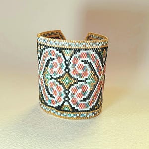 May include: A wide cuff bracelet crafted from small glass beads in a repeating geometric pattern. The design features black, white, coral, teal, and metallic gold beads arranged in symmetrical swirling shapes and diamond motifs.