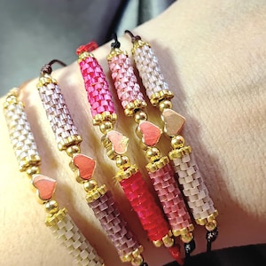 May include: A collection of beaded bracelets in various shades of pink, red, and white. Each bracelet features a cylindrical design with gold-colored accents and a heart-shaped charm. The bracelets are displayed on a wrist.