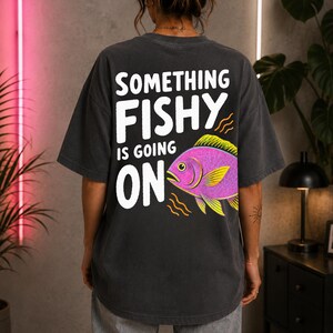 Könnte beinhalten: Ein anthrazitfarbenes T-Shirt mit dem weißen Schriftzug SOMETHING FISHY IS GOING ON auf der Rückseite. Unter dem Text befindet sich eine lebendige Illustration eines rosa Fisches mit gelben Flossen und Akzenten sowie kleinen orangefarbenen Wellenlinien.