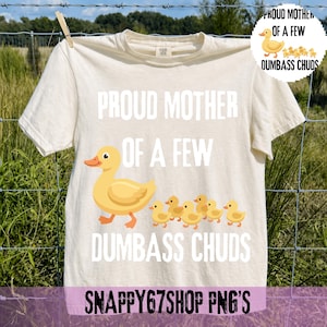 Könnte beinhalten: Cremefarbenes T-Shirt mit dem Text "PROUD MOTHER OF A FEW DUMBASS CHUDS" und einer Grafik einer gelben Ente und Küken. Das Shirt hängt an einer Wäscheleine. Der Shop-Name "SNAPPY67SHOP PNG'S" steht unten.