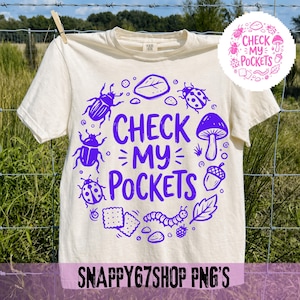 Könnte beinhalten: Cremefarbenes T-Shirt mit einem lila Grafikdesign. Das Design zeigt den Text "CHECK MY POCKETS", umgeben von Illustrationen von Insekten, einem Pilz und anderen Naturelementen. Das T-Shirt hängt an einer Wäscheleine.
