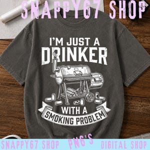 Könnte beinhalten: Ein anthrazitfarbenes Kurzarm-T-Shirt mit einem weißen Grafikdruck eines Barbecue-Smokers. Der Text lautet I'M JUST A DRINKER WITH A SMOKING PROBLEM. Mehrere Getränkedosen sind um die Basis des Grills herum abgebildet.