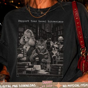 Könnte beinhalten: Ein anthrazitfarbenes T-Shirt mit einer schwarz-weißen Grafik eines Pudels, einer Katze und eines Chihuahuas in einer Bibliothek. Der Text "Support Your Local Librarians" steht über den Tieren, die in Büchern lesen.