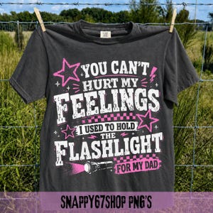 Könnte beinhalten: Dunkelgraues T-Shirt mit dem Text "YOU CAN'T HURT MY FEELINGS I USED TO HOLD THE FLASHLIGHT FOR MY DAD" in Weiß und Pink. Das T-Shirt hängt an einer Wäscheleine. Auf der Unterseite des T-Shirts steht "SNAPPY67SHOP PNG'S".