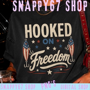 Könnte beinhalten: Ein schwarzes T-Shirt mit dem Text HOOKED ON Freedom in weißer und blauer Schrift. Das Design zeigt zwei Angelköder, die mit rot-weiß-blauen Streifen und Sternen verziert sind, umgeben von kleinen weißen, roten und blauen Sternen.