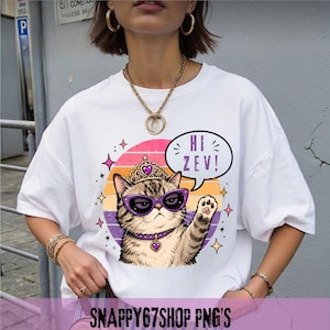 Könnte beinhalten: Weißes T-Shirt mit einer Cartoon-Katze, die eine lila Sonnenbrille und eine Krone trägt, mit dem Text "Hi Zev!" in einer Sprechblase. Die Katze ist vor einem Retro-Sonnenuntergangsdesign mit Sternen platziert.