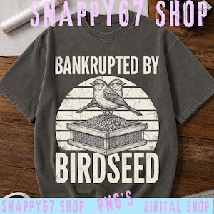 Könnte beinhalten: Ein anthrazitfarbenes Kurzarm-T-Shirt mit einem Grafikdruck von zwei Vögeln, die auf einem mit Samen gefüllten Futterhäuschen sitzen. Der Text "BANKRUPTED BY BIRDSEED" ist in weißen Großbuchstaben über und unter der Illustration gedruckt.