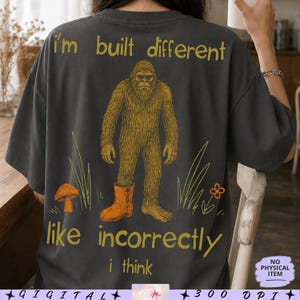 Könnte beinhalten: Ein anthrazitfarbenes T-Shirt mit einer Grafik eines braunen Bigfoot-Wesens, das einen einzelnen orangefarbenen Stiefel trägt. Der Text "i'm built different" steht oben und "like incorrectly i think" unten, ergänzt durch Pilz- und Blumenmotive.