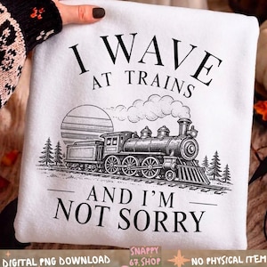 Könnte beinhalten: Ein weißes Sweatshirt mit einer schwarzen Grafik einer Dampflokomotive auf Bahngleisen, ergänzt durch eine Sonne und Tannenbäume. Der Text I WAVE AT TRAINS AND I'M NOT SORRY ist in einer fetten, schwarzen Serifenschrift abgebildet.