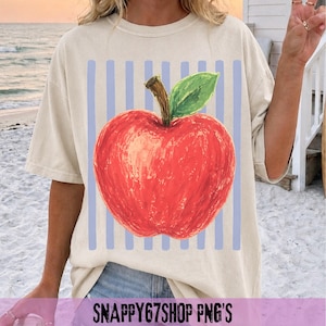 Puede incluir: Camiseta color crema con una gran ilustración de una manzana roja, una hoja verde y un tallo marrón. La manzana está sobre rayas verticales de color azul claro. El texto "SNAPPY67SHOP PNG'S" está en la parte inferior. Corte casual.