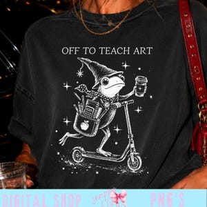 Könnte beinhalten: Ein schwarzes T-Shirt mit einer weißen Grafik eines Frosches mit spitzem Hut, der auf einem Roller fährt und einen Kaffeebecher hält. Der Frosch trägt eine Tasche mit Apfel und Stiften. Der Text OFF TO TEACH ART steht über der Illustration.