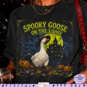 Könnte beinhalten: Ein schwarzes T-Shirt mit einer Grafik einer weißen Gans, die einen schwarz-violetten Hexenhut trägt. Der Text SPOOKY GOOSE ON THE LOOSE steht über einem gelben Mond, einer dunklen Burgsilhouette und orangefarbenen Kürbissen am Boden.