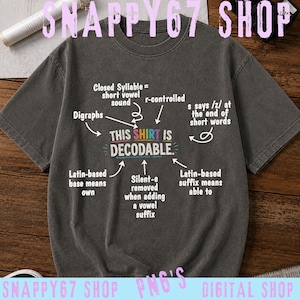Op de afbeelding: Een antracietgrijs T-shirt met korte mouwen en een afbeelding met de tekst THIS SHIRT IS DECODABLE. Het ontwerp bevat diverse pijlen die wijzen naar taalkundige uitleg zoals Closed Syllable, Digraphs, r-controlled en Latijnse achtervoegsels.