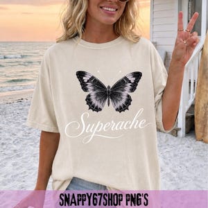 Könnte beinhalten: Cremefarbenes T-Shirt mit einem detaillierten schwarz-weißen Schmetterlingsdesign und dem Wort "Superache" in Schreibschrift. Das Shirt hat eine lockere Passform. Im Hintergrund ist eine Strand-Szene zu sehen.