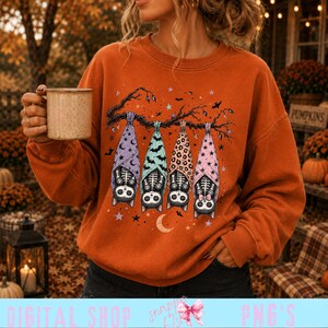 Könnte beinhalten: Ein orangefarbenes Langarm-Sweatshirt mit einer Grafik von vier Skelettkatzen, die an einem Ast hängen. Jede Katze ist in eine Decke mit unterschiedlichem Muster gewickelt, darunter violette Monde, grüne Fledermäuse, Leopardenmuster und rosa Sterne.