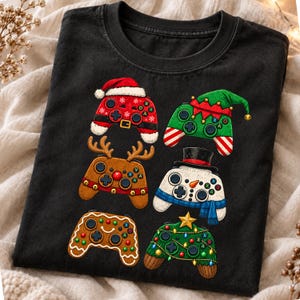 Op de afbeelding: Een zwart t-shirt met korte mouwen en een grafische print van zes voor de feestdagen versierde videogamecontrollers. De ontwerpen omvatten een kerstman-controller, een elf-controller, een rendier-controller, een sneeuwman-controller, een peperkoek-controller en een kerstboom-controller.