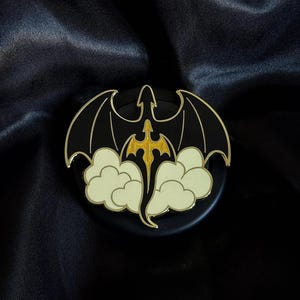 Könnte beinhalten: Schwarzer und goldener Drachen-Ansteckpin mit ausgebreiteten Flügeln, goldenem Körper und stilisiertem Wolkenmuster. Der Pin hat einen goldenen Rand und ist vor einem dunklen Hintergrund platziert.