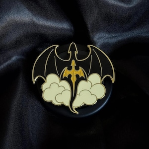 May include: A black and gold dragon pin with outstretched wings, a gold body, and cream-colored clouds. The pin has a gold border and is set against a dark background.